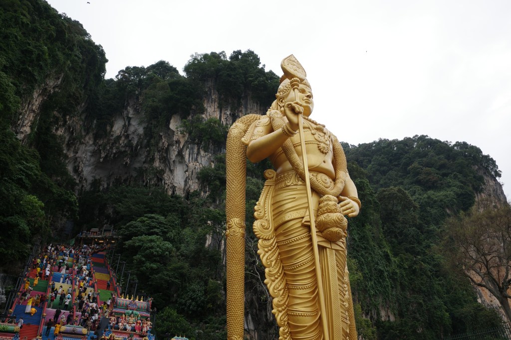 A large gold Hindu statue stands in front of a steep mountainside with colorful stairs full of people leading to the Batu Cave entrance at the top.