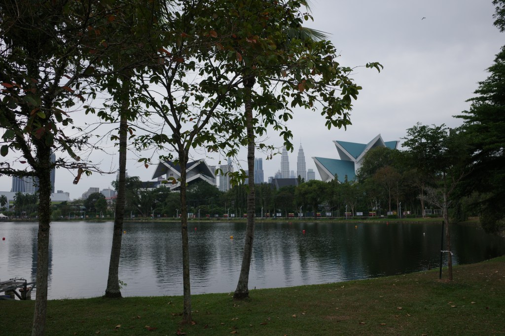 A view of Kuala Lumpur's Chinatown buildings, shot from a small lake with trees in the foreground.