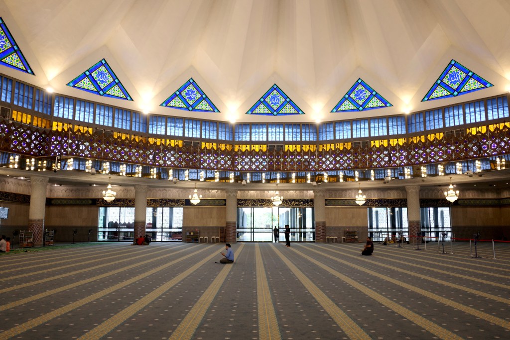 The interior of a mosque in Kuala Lumpur with a few people kneeling on the carpeted floor and a domed white ceiling with graphic stained glass windows.