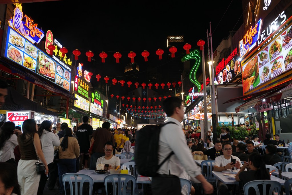 A busy Kuala Lumpur Chinatown street scene with vendors, people eating and colorful lights.