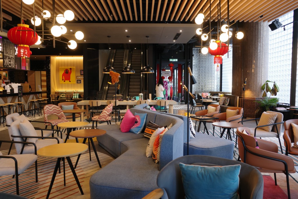 The colorful lobby area of the Moxy Kuala Lumpur Chinatown with a bar at the far left, wavy blue couches, small round tables, graphic artwork on the walls and two escalators leading upstairs.