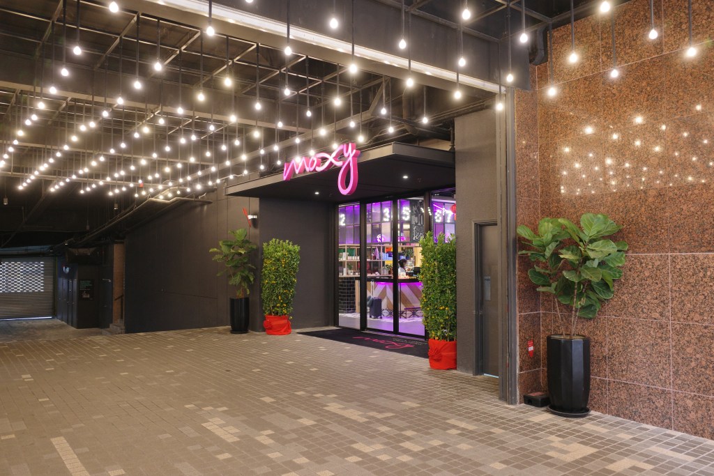 The entrance to the Moxy Kuala Lumpur Chinatown with a pink neon sign, lights overhead and tropical plants flanking the doors. 