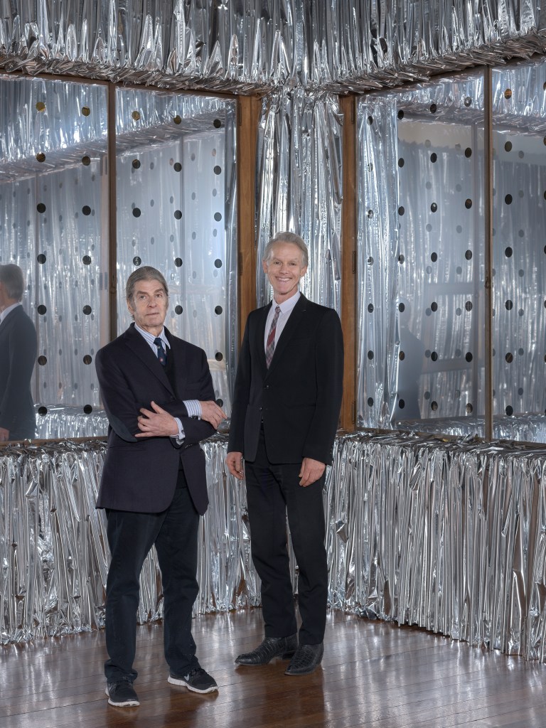 Marc-Olivier Wahler and John Armleder stand in front of a silver and plastic abstract art installation.