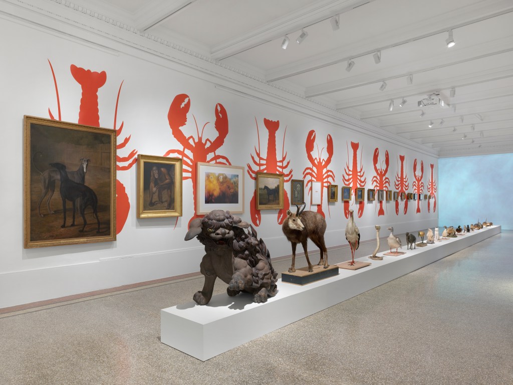 A long, white art gallery room with a row of framed animal painting handing on red lobsters paintings on the wall, and a row of animal sculptures of various sizes. 
