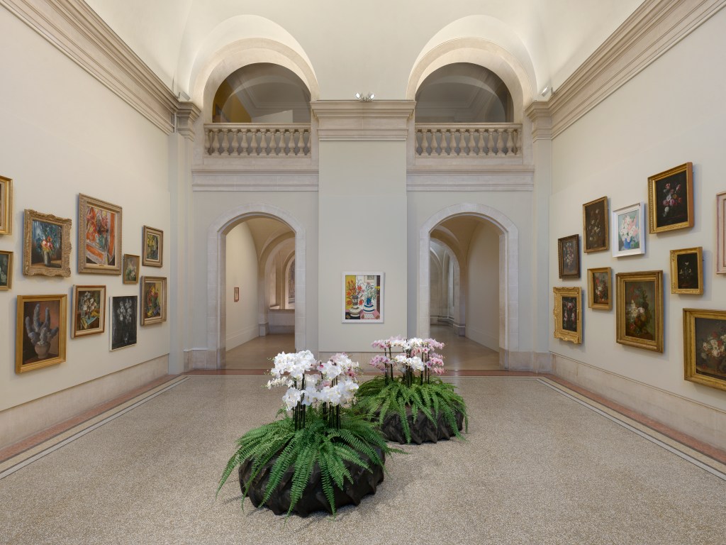 A gallery room at MAH Genève wtih two aches doorways, a balcony, two large potted plants and several paintings hung on both walls flanking the walkway.