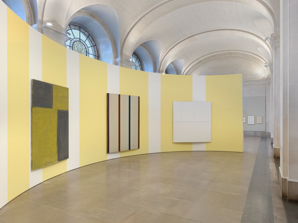 A curved yellow and white painted wall with three minimal abstract paintings hanging on it in a museum gallery room with tall arched ceiling panels. 