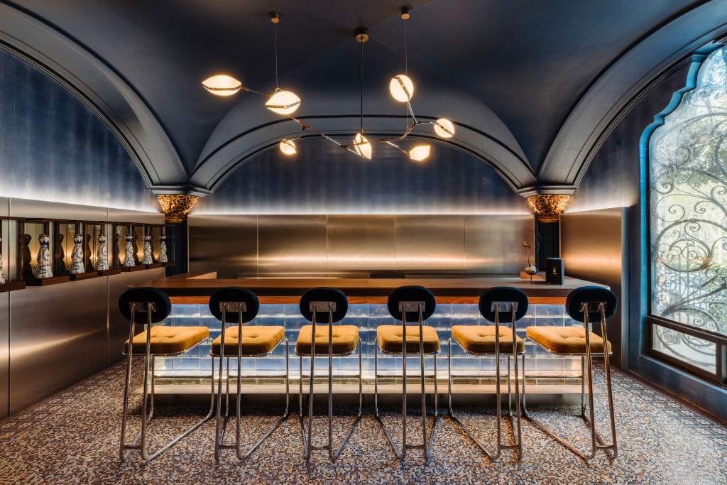 A gray domed ceiling room with a wooden and glass bar at the center, an asymmetrical chandelier and a large ornate window on the right side.