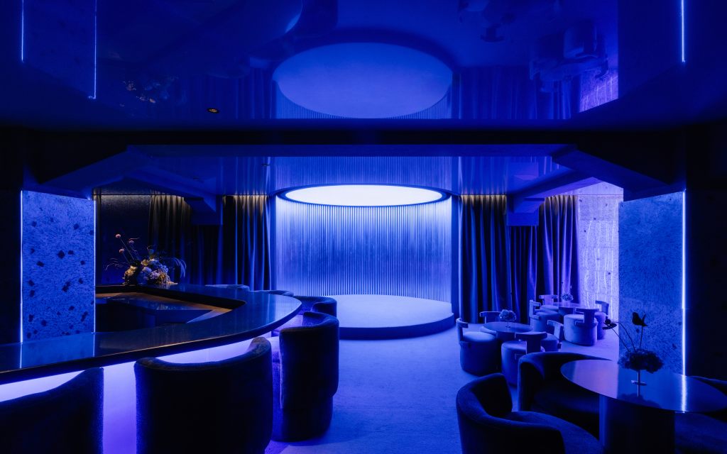 A blue room at Casa de los Leones featuring a curved bar, small round cocktail tables with architectural chairs and a central stage area. 