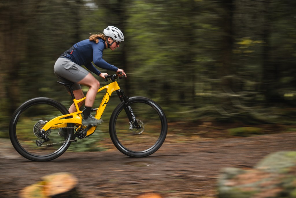 A side view of a person with long hair riding a yellow mountain bike on a wooded trail.