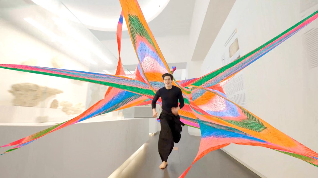 A man wearing black runs through a museum trailed by brightly colored shapes streaming behind him.