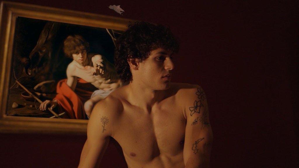 A man without a shirt looks to the right of frame as a saint in the painting behind him reaches towards him.