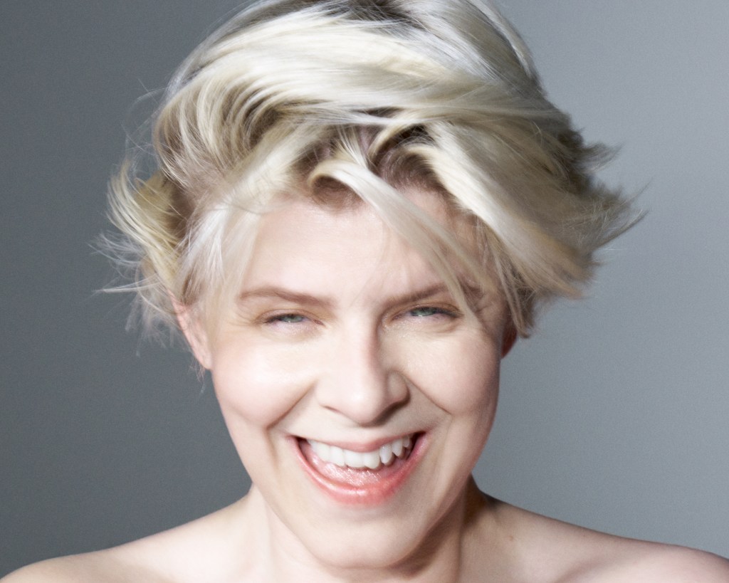 A photo of Robyn with blonde short hair, bare shoulders and a large smile, shot on gray background.