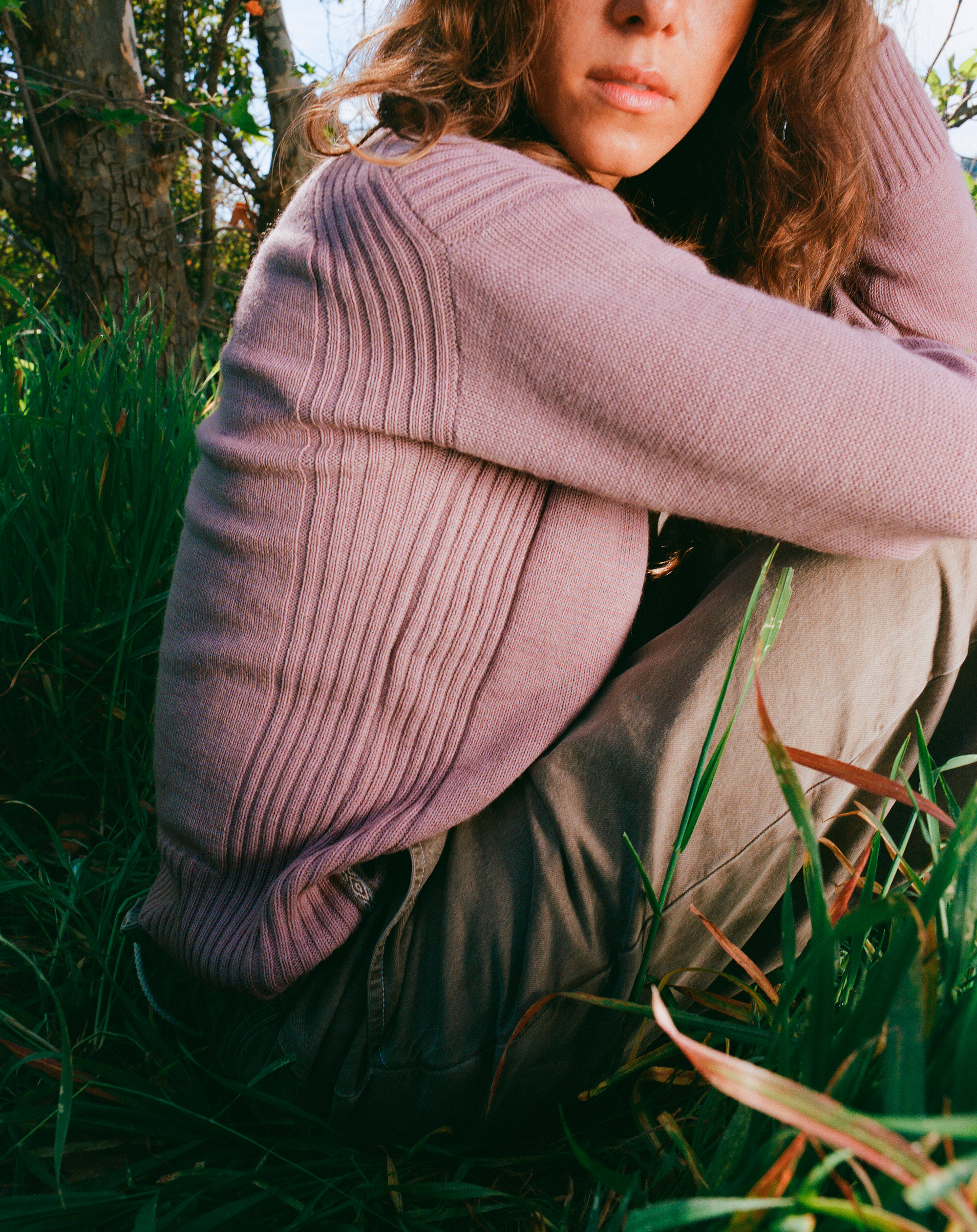 A tight crop of a woman wearing a pink alpaca PAKA sweater, crouding down with one arm resting on her leg, one arm holding her head.