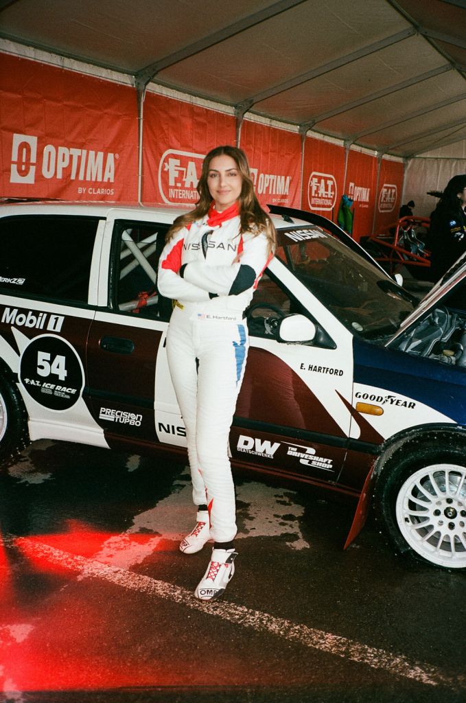 A racecar driver wearing a white uniform standing in front of her Mobil 1 team car with a red banner in the background.