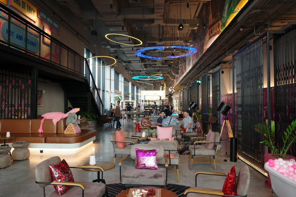 The hotel lobby area at the Moxy Bangkok with gray chairs, colorful and sequined pillows, people sitting and a row of pink, numbered lockers on the right side of the frame. 