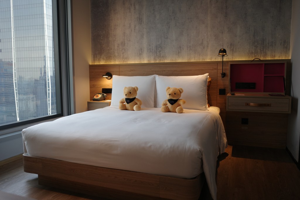 A double bed with teddy bears and a light wood headboard in a guest room at Moxy Bangkok.