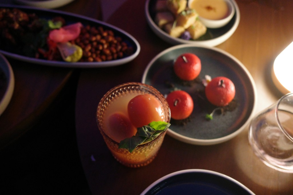 A dimly lit wood table with an orange cocktail and bowls of Thai snacks, shot overhead.