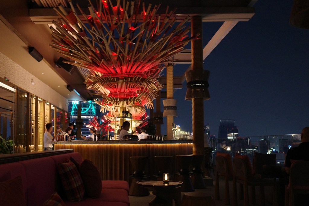 The rooftop bar at the Moxy Bangkok with a large sculptural bamboo ceiling and tables overlooking the city at night.