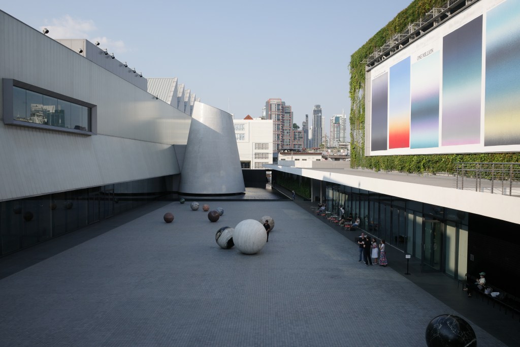The sculptural exterior of Dib Bangkok with a courtyard containing large spherical sculptures, people loitering nearby, and a large green plant wall with a billboard of colorful gradients that says "ONE MILLION."