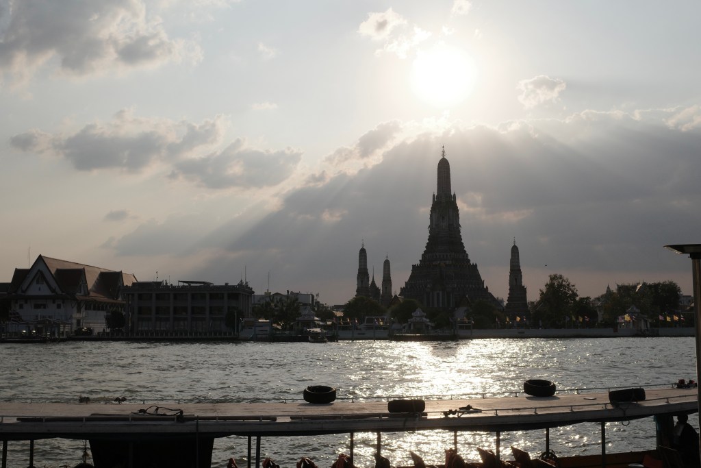 The Erawan and Trimurti Shrines, shot across a large reflective body of water, with high sun above.