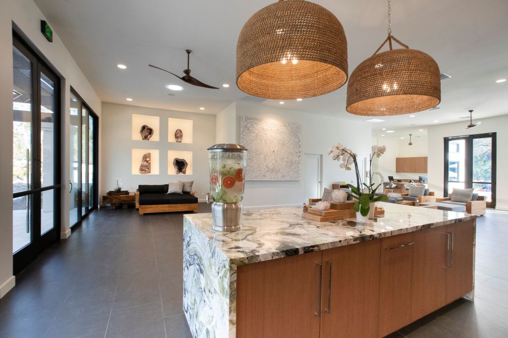 The light-filled lobby area of We Care Spa with a seating area in the corner, large wicker light fixtures hanging over the kitchen island, and a self-serve juice bar for guests.