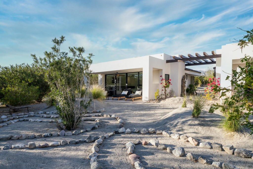 The white stucco exterior and pergola roof of a suite at We Care Spa, surrounded by stones and desert plants.