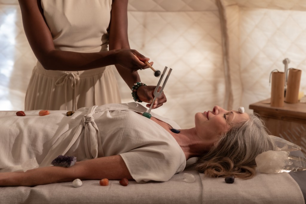 A woman lying on a table with crystals on her chakras getting a spa treatment by a practitioner who is holding a tuning fork.