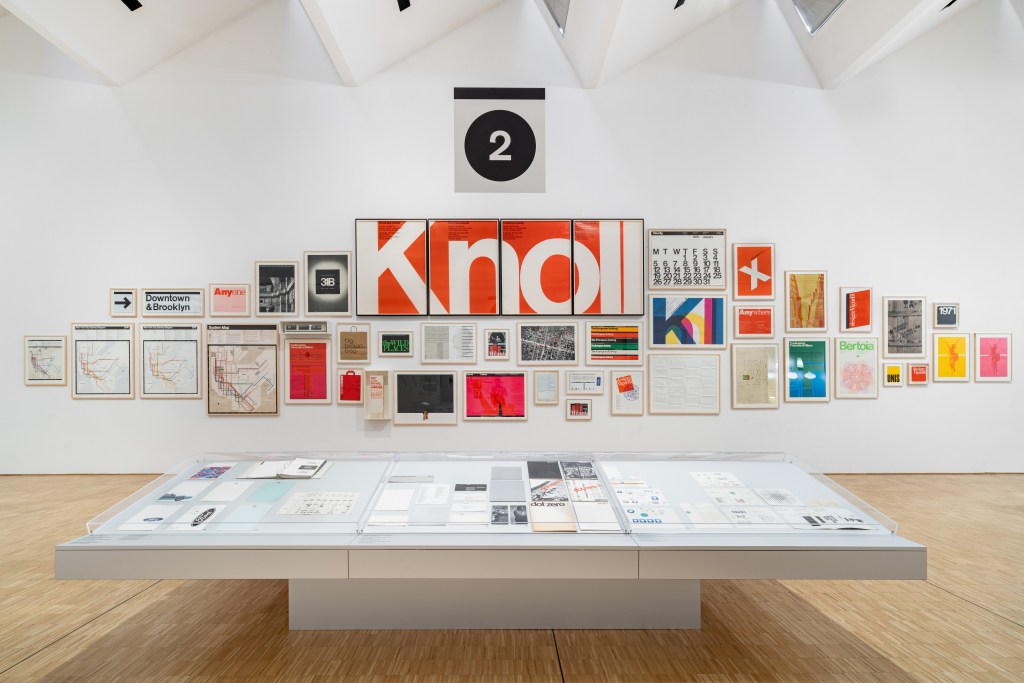A white wall of framed Knoll graphic design examples by Lella and Massimo Vignelli with a light wood table of other designs in front.
