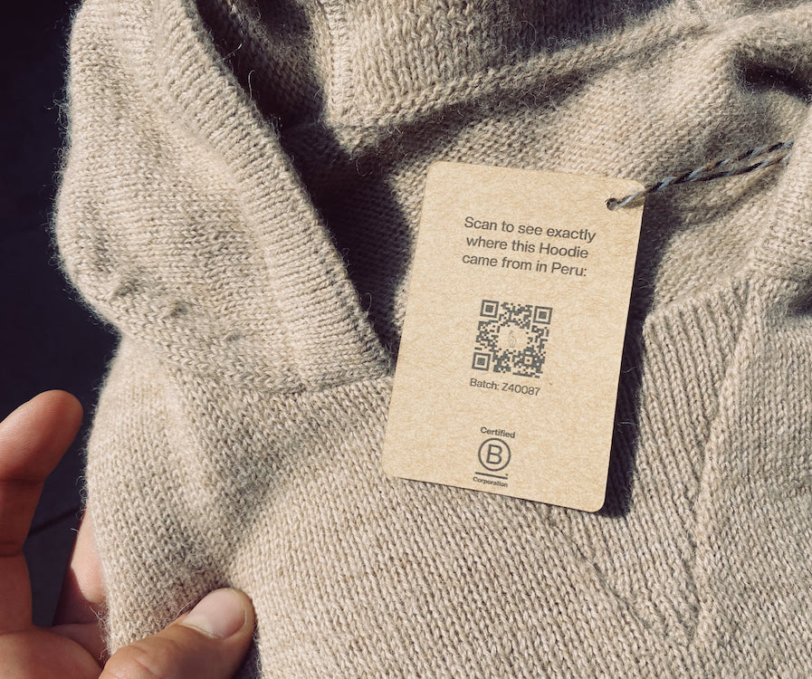 A detail of an alpaca PAKA hoodie with a brown kraft paper tag that says "Scan to see exactly where this Hoodie came from in Peru: with a QR code below.