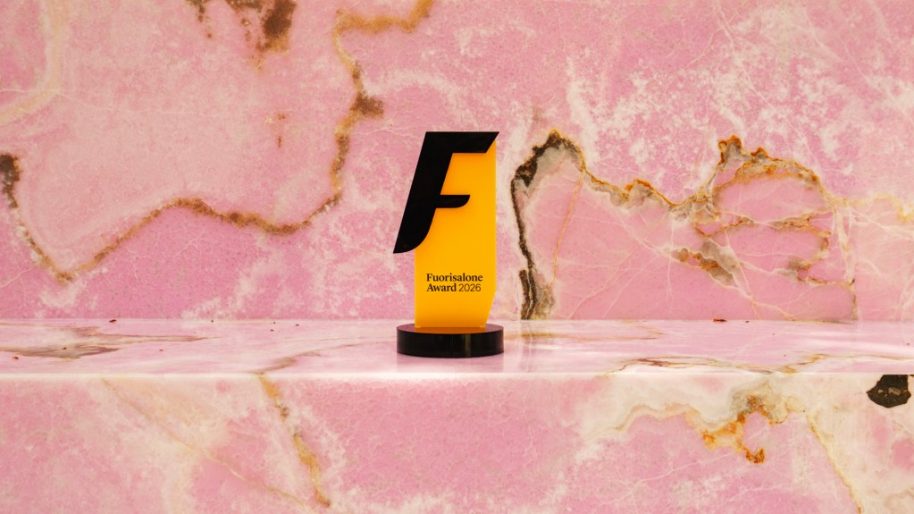 The Fuorisalone Award 2026, a golden yellow award with a black "F" on it, shot on a pink stone surface.