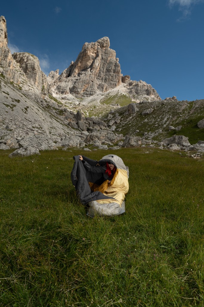 A person wrapping themselves into The North Face Universal One Bag on a grassy surface with a mountain in the background.
