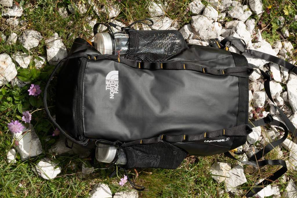 A black The North Face backpack lying on a grassy and rocky surface.