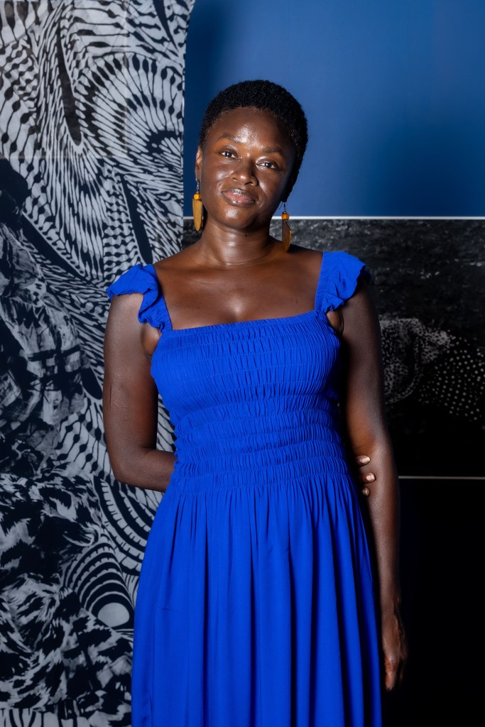 Keisha Scarville stands in a bright blue dress in front of her black-and-white artwork.