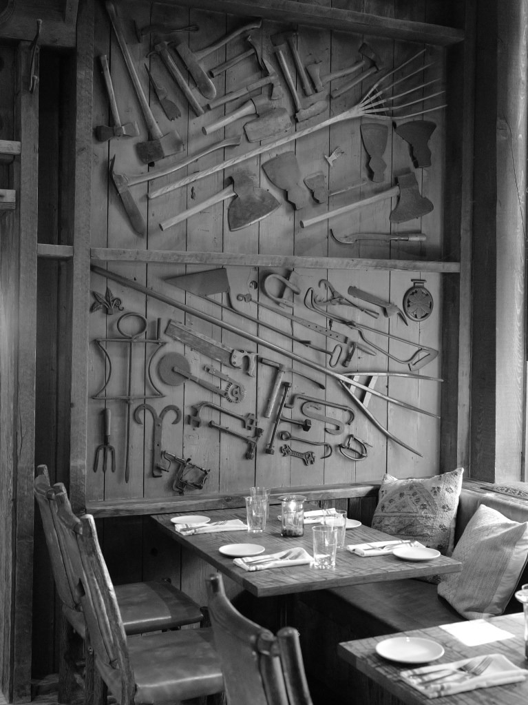 A black and white photo of a wall in The Foundry restaurant with antique farming equipment hanging on the wall above a dining table.