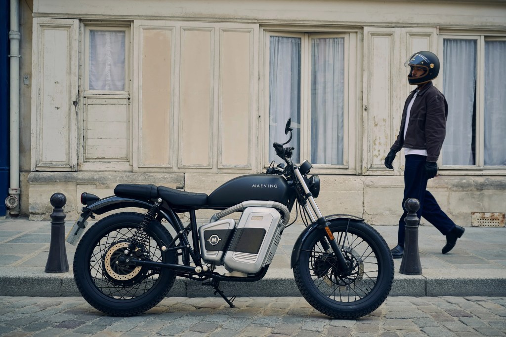 A Maeving RM2 electric motorcycle parked on a city street in front of a cream building with a rider walking toward it fro the right side of frame.