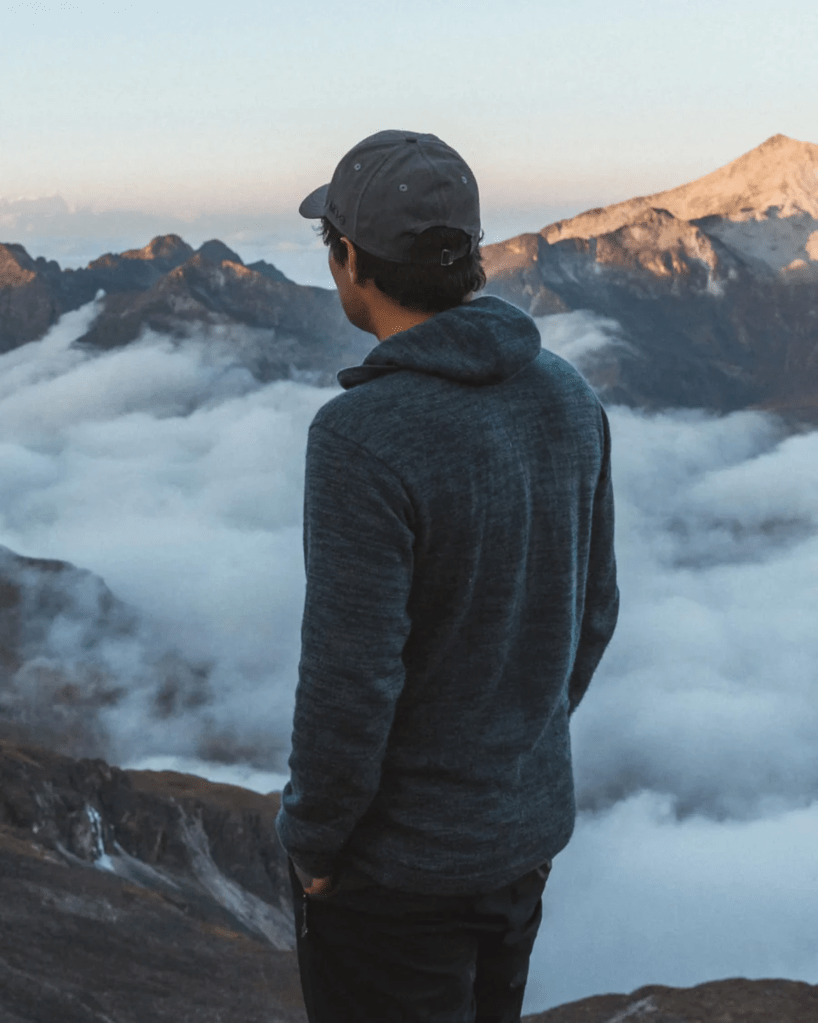 A man wearing an alpaca PAKA hoodie looks out onto the clouds hovering over a mountain range.
