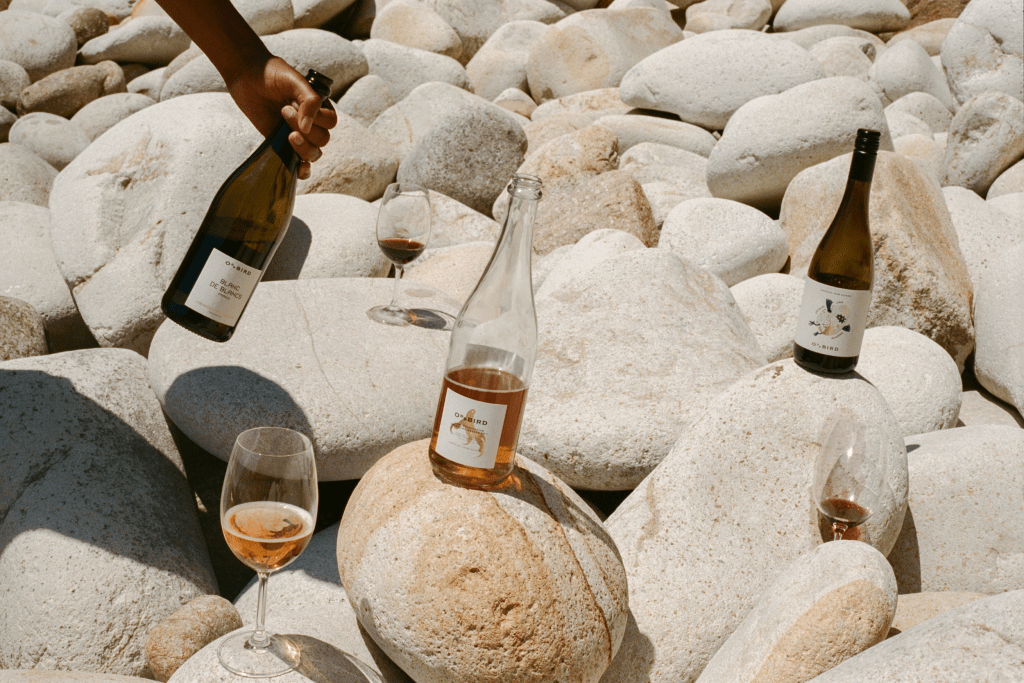A group of large white rocks with two non-alcoholic wine bottles on wine of the rocks, three wine glasses with wine and one hand on the left side of the frame holding a bottle.