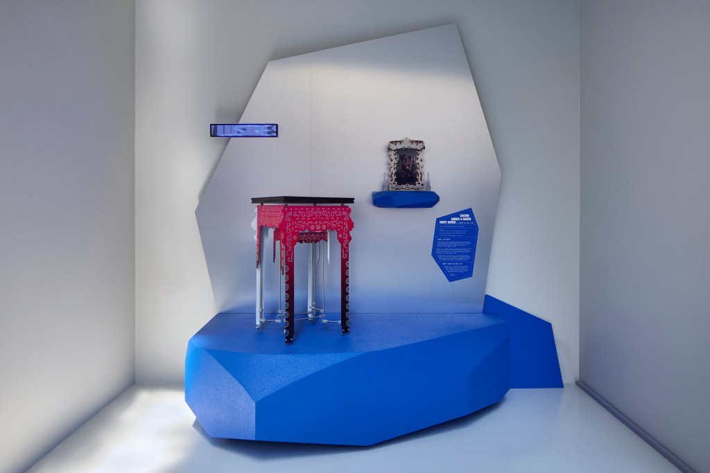 An exhibit at Prototype Island with a blue geometric base, silver panel, an ornate pink table, a small horizontal digital screen and an ornate small frame.