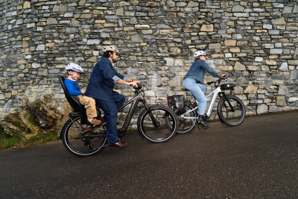 A man and a woman riding Vado EVO bikes uphill with a small child riding on the back of the man's bike.
