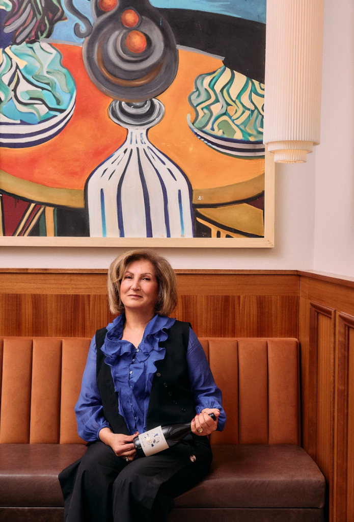 Oddbird founder Moa Gürbüzer wearing a blue top and black vest while holding a bottle of Oddbird wine, sitting on a brown leather banquette in front of a colorful painting.