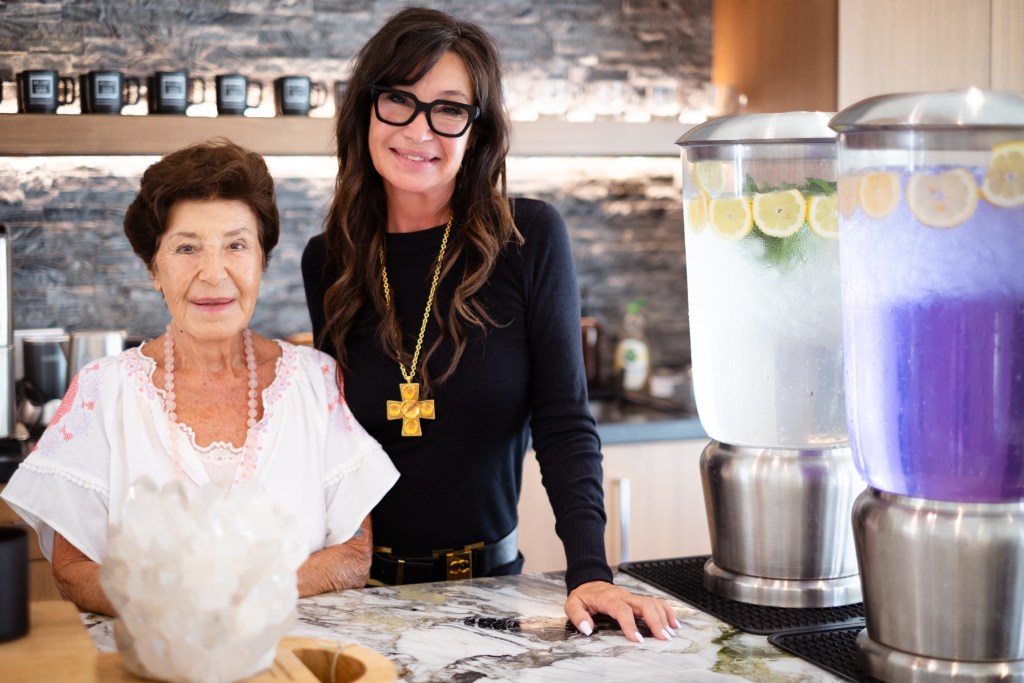 Susan Belen and her daughter Susan Lombardi, standing next to each other by tall carafes of fruit waters in the lobby of We Care Spa.