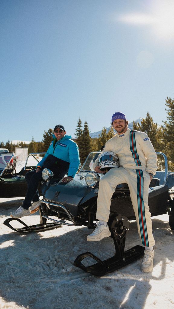 Two racers sitting on the hood of a car on a snowy track with evergreens in the background.