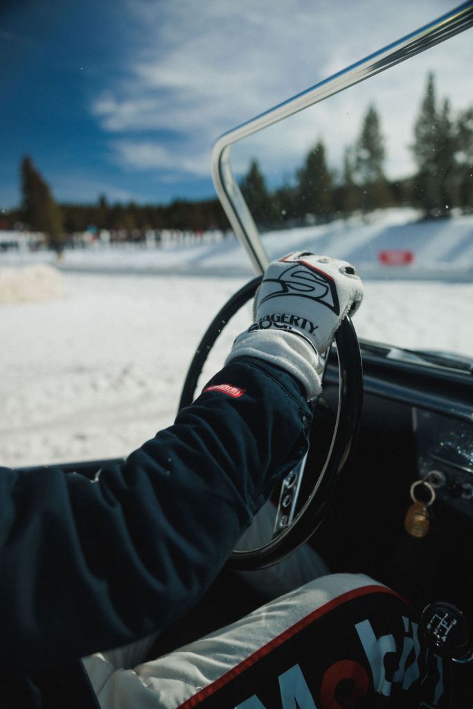 A driver's arm on a steering wheel with snow and evergreen trees in the background.