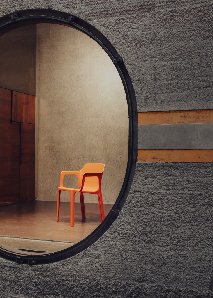 An orange Linnea chair, seen as a reflection in a circular mirror hanging on a textured concrete wall.