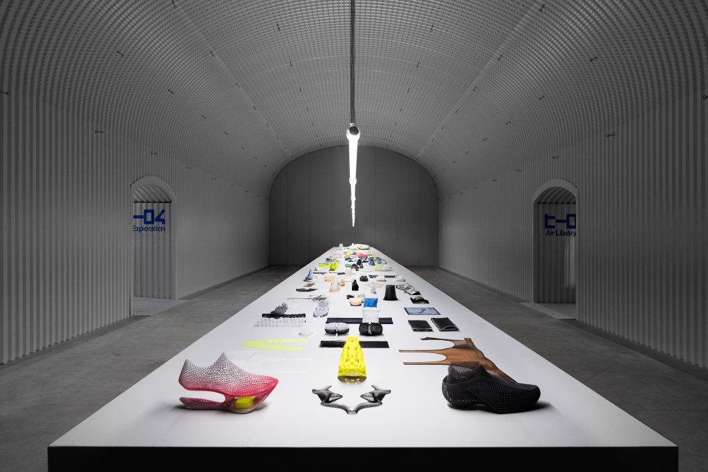 A light gray room with a long table at the NikeAir_Lab installation, showing a variety of colorful Nike technological components laid out in an organized way on the white surface, flanked by two arched doorways.