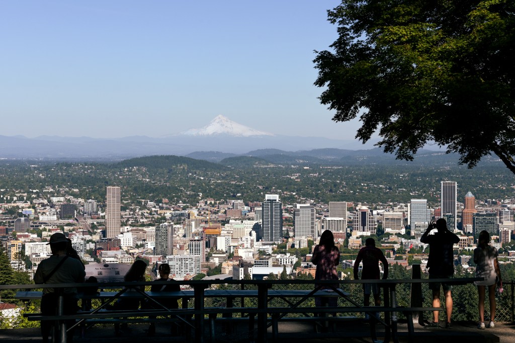 The backs of several people standing on a lookout point, looking out onto a view of downtown Portland with Mt Hood in the background.