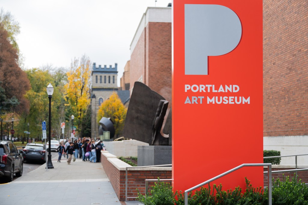An exterior view of a red sign with a large gray "P" that reads "Portland Art Museum" below it, with a group of children in the background.