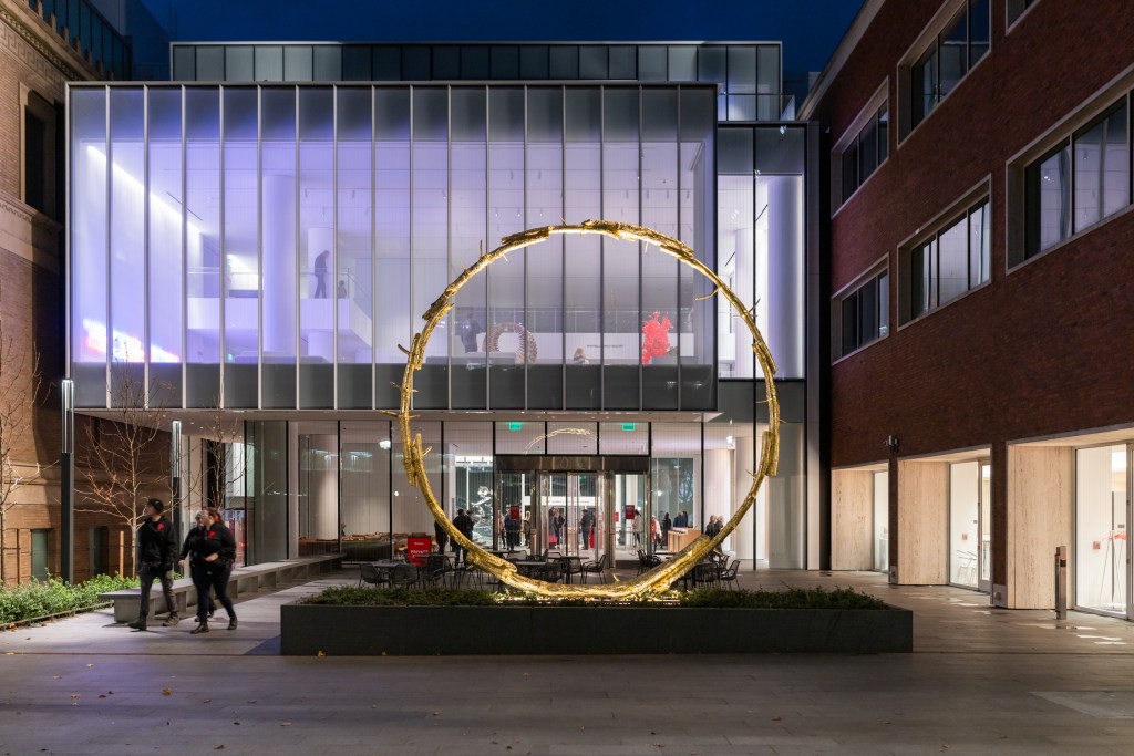 A nighttime shot of the Portland Art Museum with a large, illuminated gold ring sculpture in front of the entrance.