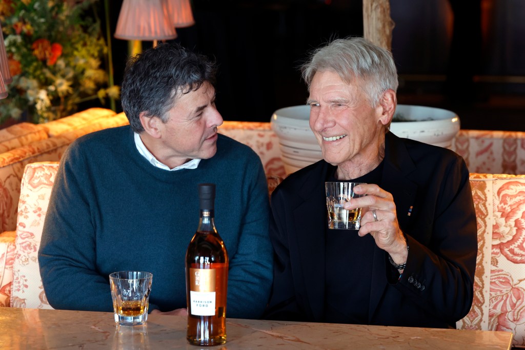 Dr. Bill Lumsden and Harrison Ford sitting next to each other, behind a wooden table and a bottle of their limited edition Glenmorangie single malt scotch.