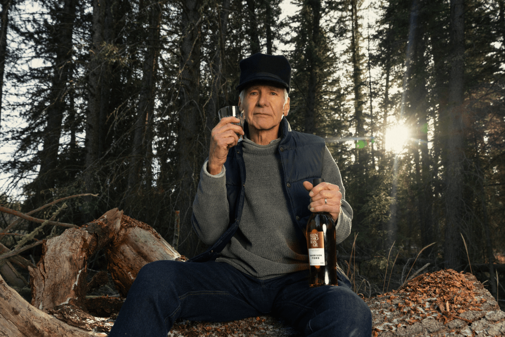 Harrison Ford sitting on a large rock outside in the woods, holding a bottle of his Glenmorangie single malt whisky with tall trees behind him.